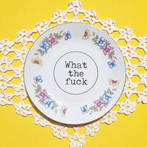 Può includere: Piatto in ceramica bianca con disegni floreali e farfalle, e la scritta "What the fuck" al centro. Il piatto è su una tovaglietta di pizzo bianco, su sfondo giallo.
