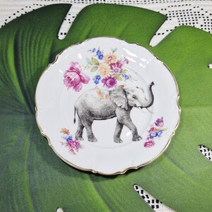 Könnte beinhalten: Weiße Keramikplatte mit goldenem Rand, mit einer grauen Elefantenillustration und floralen Akzenten in Rosa, Blau und Gelb. Der Teller hat einen gewellten Rand.
