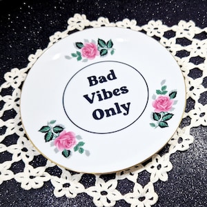 Può includere: Piatto in ceramica bianca con bordo dorato e una grafica in bianco e nero che dice "Bad Vibes Only". Il piatto è decorato con rose rosa.