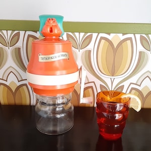 May include: Orange and white ice crusher with a cartoon crab on top. The crusher has a label that reads "Tritaghiaccio ad Pirata". A glass of orange juice with a lemon wedge is in the foreground.