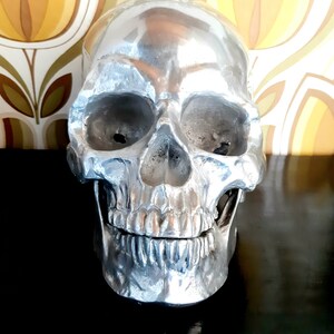 May include: A silver skull sculpture with a detailed, textured surface. The skull has a realistic appearance with a defined jawline and teeth.