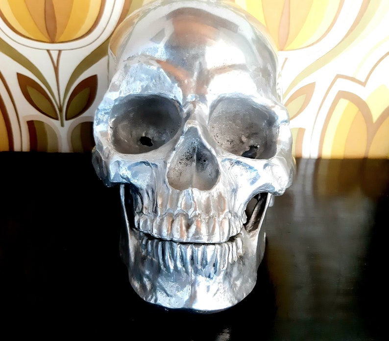 May include: A silver skull sculpture with a detailed, textured surface. The skull has a realistic appearance with a defined jawline and teeth.