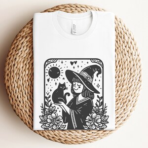 Peut inclure: T-shirt blanc avec une illustration en noir et blanc d'une sorci&egrave;re tenant un chat noir. Le motif comprend un chapeau de sorci&egrave;re, des fleurs, un soleil et des &eacute;toiles, cr&eacute;ant un graphisme fantaisiste sur le th&egrave;me d'Halloween.