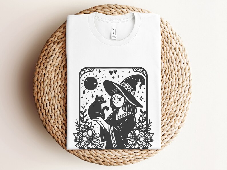 Peut inclure: T-shirt blanc avec une illustration en noir et blanc d'une sorci&egrave;re tenant un chat noir. Le motif comprend un chapeau de sorci&egrave;re, des fleurs, un soleil et des &eacute;toiles, cr&eacute;ant un graphisme fantaisiste sur le th&egrave;me d'Halloween.