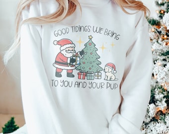 May include: White sweatshirt with a Christmas-themed graphic featuring Santa Claus, a Christmas tree, and a dog. The text reads "Good Tidings We Bring To You And Your Pup."