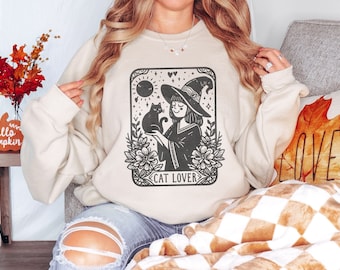 May include: Cream-coloured sweatshirt featuring a black graphic of a witch with a black cat, framed by flowers and celestial symbols. The words "CAT LOVER" are below. The top is a neutral colour, ideal for autumn or Halloween.