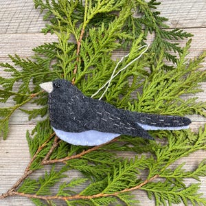 May include: A felt bird ornament with a gray body, white belly, and black eyes. The ornament is hanging from a branch of a green evergreen tree.