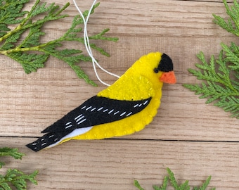 May include: A felt ornament of a yellow bird with black wings and a red beak. The bird is hanging from a white string.