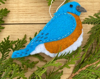 May include: A bluebird ornament made of felt with a white belly and orange breast. The bird has a black eye and a black beak. The ornament is hanging from a white string.