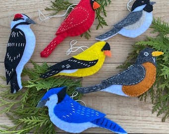 May include: Five felt bird ornaments, each with a different design and colour. The birds are a cardinal, a woodpecker, a blue jay, a goldfinch, and a robin.