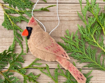 May include: A felt cardinal ornament with a red beak and black eyes. The ornament is hanging from a white string and is surrounded by green cedar branches.