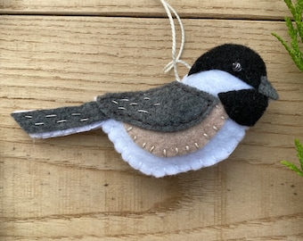 May include: A felt bird ornament with a grey body, white belly, and black head. The bird has a small, black eye and a white string for hanging.