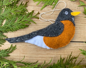 Puede incluir: Un adorno de robin hecho de fieltro con cabeza gris, pecho naranja y vientre blanco. El robin tiene un ojo negro y un pico amarillo. El adorno está colgado de una cuerda blanca.