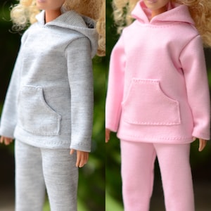 May include: Two doll outfits: a grey hoodie and matching trousers, and a pink hoodie and matching trousers. Both outfits have a front pocket on the hoodie and are made of a soft fabric. The outfits are designed for dolls.