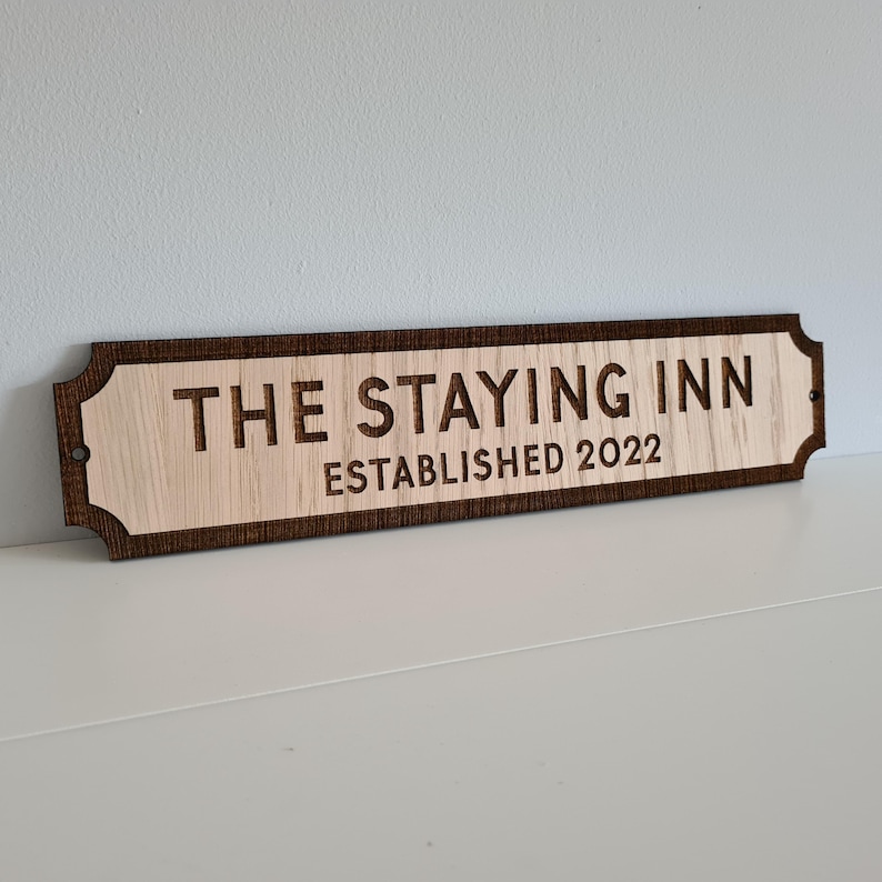 May include: A wooden sign with the text "The Staying Inn Established 2022" in a brown font. The sign has a dark brown border and rounded edges.
