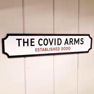 May include: A white sign with black trim and rounded corners. The sign reads "THE COVID ARMS ESTABLISHED 2020".