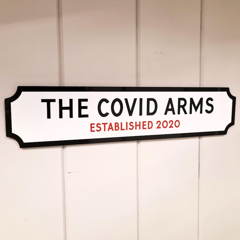 May include: A white sign with black trim and rounded corners. The sign reads "THE COVID ARMS ESTABLISHED 2020".