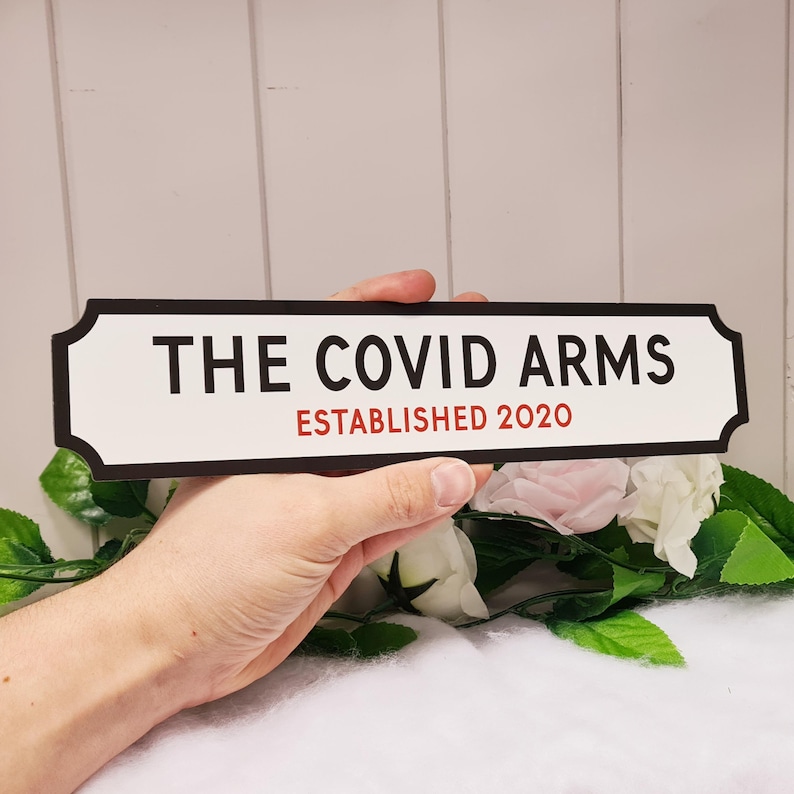 May include: A white sign with black lettering that reads "The Covid Arms Established 2020". The sign is held in a hand.