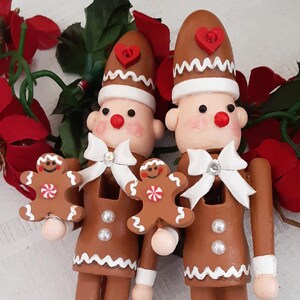 May include: Two wooden Christmas figurines of gingerbread men. They have brown bodies, white bow ties, and hats with red heart accents. Each holds a gingerbread man cookie. The figurines stand on white bases.