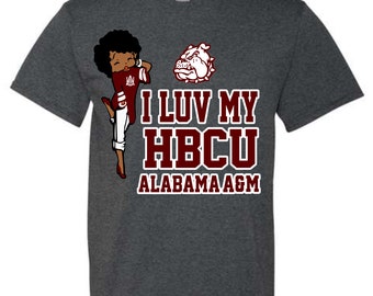May include: A dark heather grey t-shirt with a cartoon image of a woman wearing a maroon and white uniform with a bulldog mascot. The text "I Luv My HBCU Alabama A&M" is printed in maroon on the shirt.