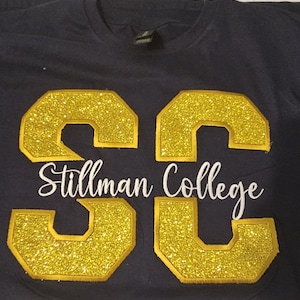 May include: A navy blue t-shirt with a white embroidered design that says "Stillman College" and two large gold glitter letters "SC".