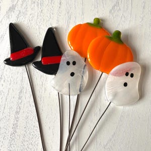 May include: A selection of Halloween garden stakes made of glass. The stakes depict black witch hats with red bands, orange pumpkins, and white ghosts with black dot eyes. They are mounted on thin, dark metal rods.