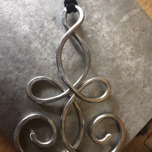 May include: A silver pendant necklace with a unique, intricate design. The pendant is shaped like a stylized infinity symbol with multiple loops and swirls. The necklace is made of metal and has a black cord.