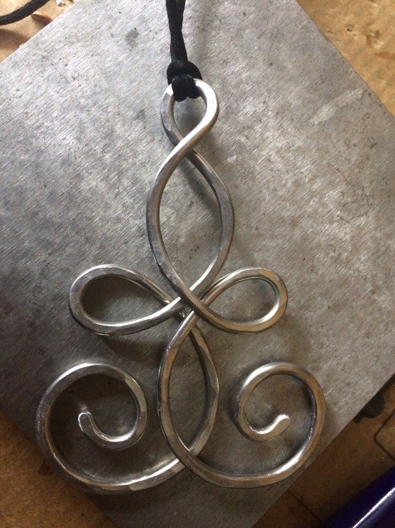 May include: A silver pendant necklace with a unique, intricate design. The pendant is shaped like a stylized infinity symbol with multiple loops and swirls. The necklace is made of metal and has a black cord.
