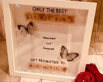 May include: White shadow box frame with a white background and a clear front. The frame contains the words "ONLY THE BEST" and "S.I.S.T.E.R.S." spelled out with wooden letter tiles. There are two butterflies, one white and one black, and a 5x5 inch photo space. The bottom of the frame contains the words "GET PROMOTED TO" and "A.U.N.T.I.E." spelled out with wooden letter tiles. The frame is sitting on a wooden surface.