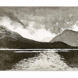 May include: A black and white print of a mountain range reflected in a still lake. The sky is cloudy and the mountains are silhouetted against the light.