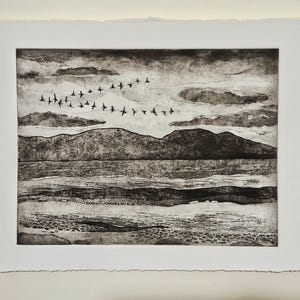 May include: Monochrome landscape print depicting a flock of birds flying over water and hills. The artwork is framed with a white border and has a textured appearance. The sky is filled with clouds, creating a dramatic scene.