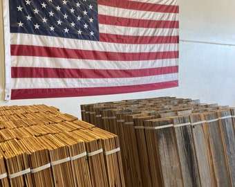 May include: A stack of reclaimed wood planks, some are light brown and some are dark brown with gray streaks. The planks are bundled together with white rubber bands. An American flag hangs on the wall behind the wood planks.