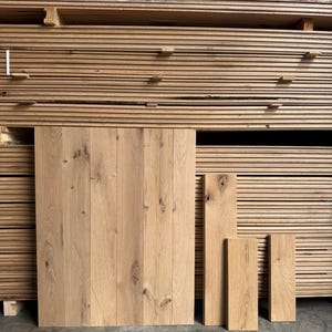 May include: A collection of light brown oak wood planks featuring visible natural grain patterns and knots. The planks are arranged in a vertical stack, with several individual boards standing upright in the foreground, showcasing the texture.
