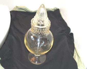 May include: A clear glass jar with a decorative lid. The jar has a round body and a pedestal base. The lid is cone-shaped and has a textured surface. The jar is likely for storage or display.