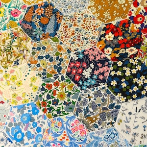 Op de afbeelding: Een patchwork van zeshoekige stofstukken. De stoffen tonen verschillende bloempatronen in tinten blauw, geel, rood en groen. De opstelling creëert een kleurrijk en gestructureerd oppervlak.