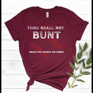 Puede incluir: Camiseta granate con texto blanco que dice "Thou Shall Not Bunt" con "Bunt" tachado. Debajo del texto está "Unless You Can Beat The Throw". Diseño de costura de béisbol en rojo.