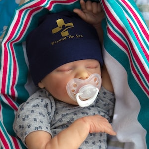May include: A sleeping baby wearing a blue hat with the text "Beyond The Sea" and a white pacifier. The baby is wearing a gray shirt with a white diaper and is lying on a striped blanket.