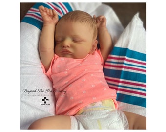 May include: A sleeping reborn baby doll wearing a pink shirt with white "XO" print and a white diaper. The doll is lying on a white and blue striped blanket.