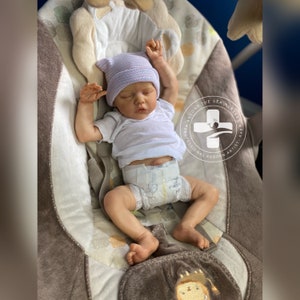May include: A realistic baby doll wearing a white shirt, a blue and white striped hat, and a diaper. The doll is lying in a brown and white patterned baby swing. The swing has a white logo that says "Beyond the Sea Nursery - Professional Reborn Artist - Rae".