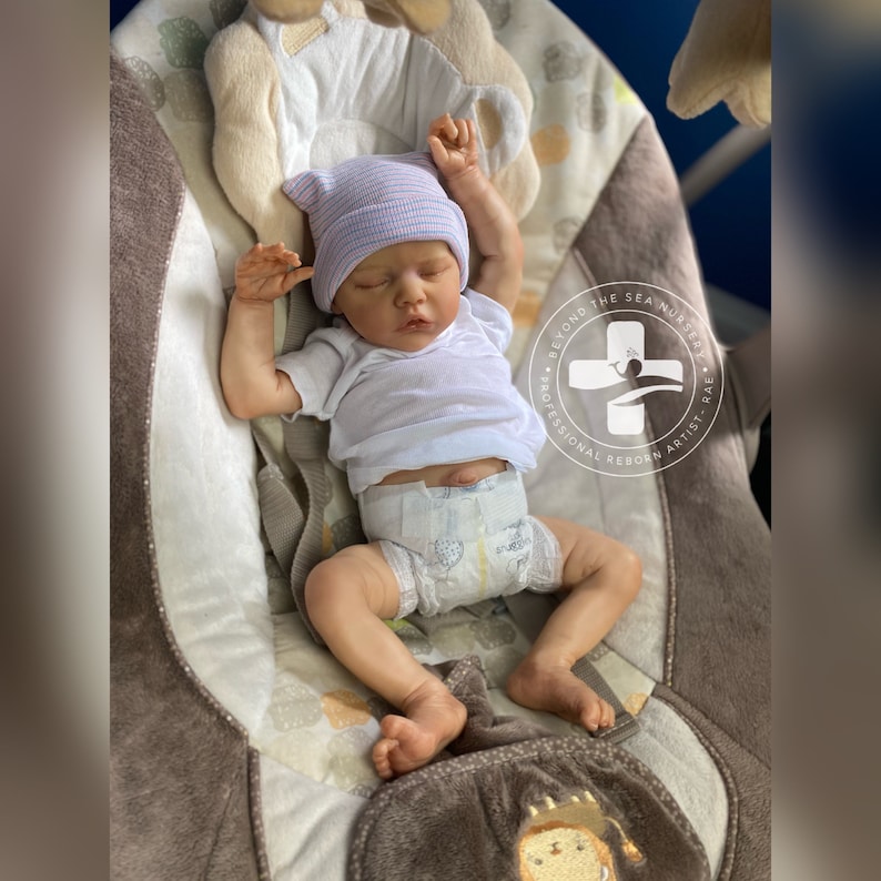 May include: A realistic baby doll wearing a white shirt, a blue and white striped hat, and a diaper. The doll is lying in a brown and white patterned baby swing. The swing has a white logo that says "Beyond the Sea Nursery - Professional Reborn Artist - Rae".
