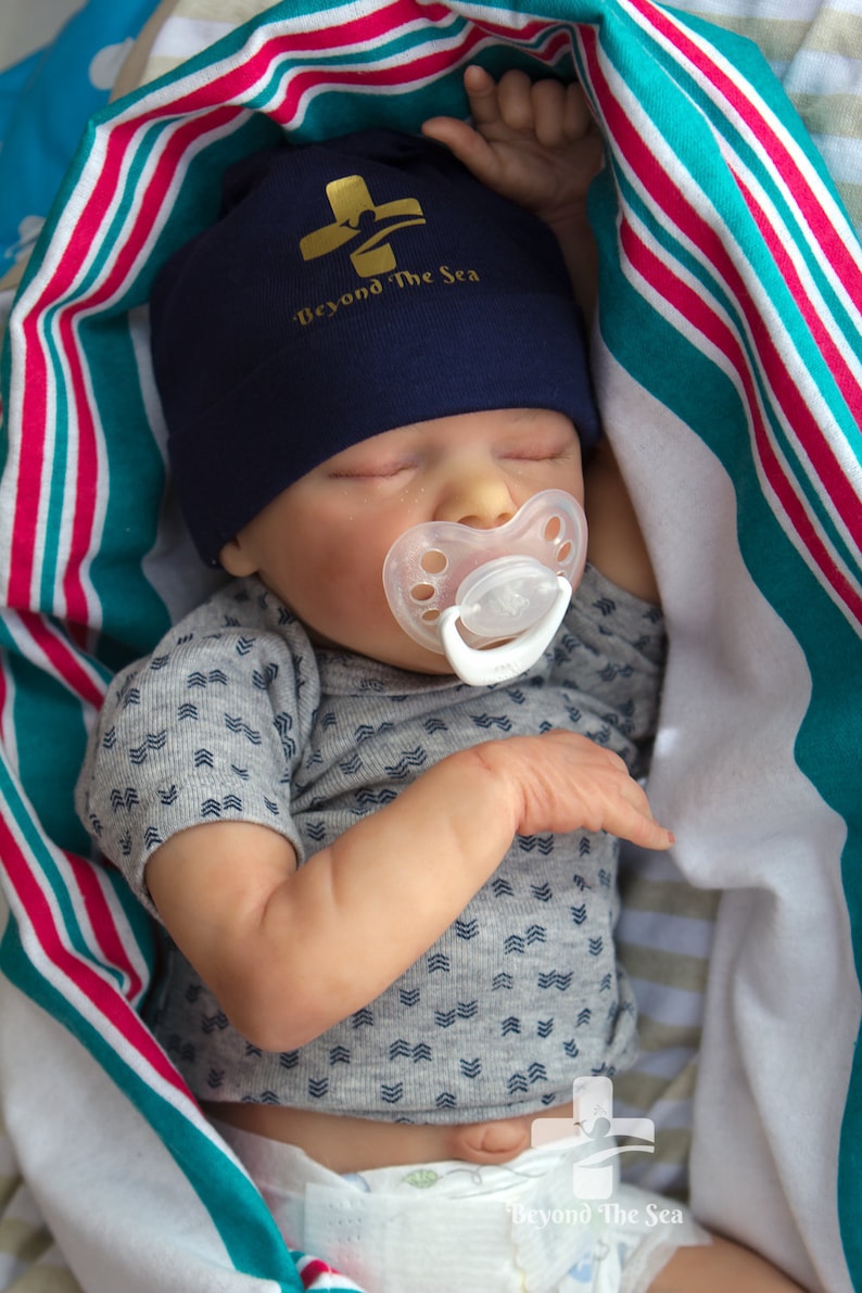 May include: A sleeping baby wearing a blue hat with the text "Beyond The Sea" and a white pacifier. The baby is wearing a gray shirt with a white diaper and is lying on a striped blanket.
