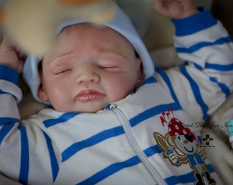 May include: A sleeping baby doll wearing a blue and white striped outfit with a pirate patch on the sleeve. The doll is wearing a blue and white hat and has a peaceful expression on its face.
