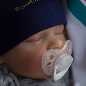 May include: A sleeping baby wearing a navy blue beanie with a gold cross and the text "Beyond The Sea". The baby is wearing a gray and white patterned onesie and has a white pacifier in their mouth.