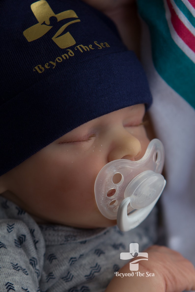 May include: A sleeping baby wearing a navy blue beanie with a gold cross and the text "Beyond The Sea". The baby is wearing a gray and white patterned onesie and has a white pacifier in their mouth.