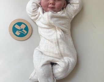 May include: A realistic newborn doll with a smooth, pale complexion, eyes closed, and arms raised above its head. The doll wears a long-sleeved, cream-colored ribbed footed sleeper with a front zipper. Beside it lies a round wooden disc with a teal cross.