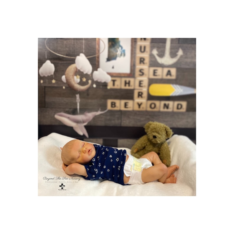 May include: A sleeping baby doll wearing a blue and white star patterned shirt and a diaper. The doll is lying on a white blanket with a teddy bear nearby. The background features a wooden wall with a nursery rhyme decoration.