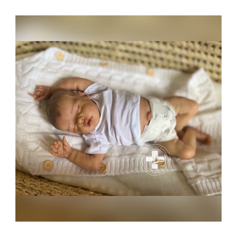 May include: A sleeping baby doll wearing a white t-shirt and a diaper with a white and gray pattern. The doll is lying on a white knitted blanket.
