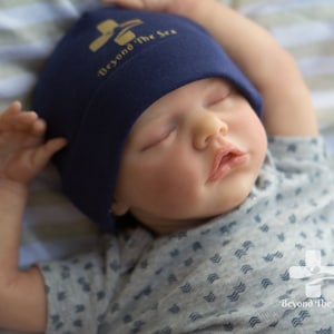 May include: A sleeping baby wearing a navy blue beanie with the text "Beyond The Sea" in gold lettering and a gray and white patterned shirt with the same logo on the bottom.