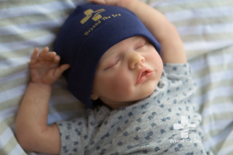 May include: A sleeping baby wearing a navy blue beanie with the text "Beyond The Sea" in gold lettering and a gray and white patterned shirt with the same logo on the bottom.