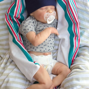 May include: A sleeping baby doll wearing a blue and gold hat with the text "Beyond the Sea" and a white pacifier. The doll is wrapped in a striped blanket with red, white, and teal stripes.